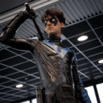 A cosplayer dressed as Nightwing poses confidently, gripping a custom 3D-printed Escrima stick. The black and blue armored suit, tactical gear, and signature mask create a powerful, heroic look.
