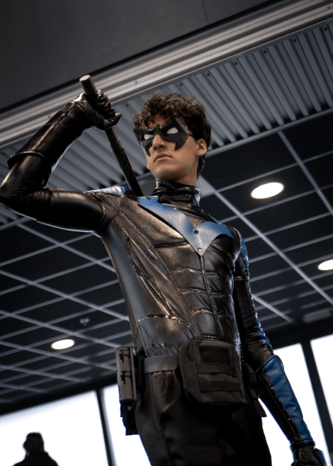 A cosplayer dressed as Nightwing poses confidently, gripping a custom 3D-printed Escrima stick. The black and blue armored suit, tactical gear, and signature mask create a powerful, heroic look.