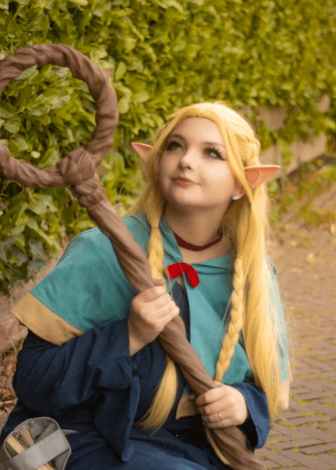 A cosplayer portrays Marcille, the elven mage, in a stunning fantasy costume. She holds a custom-printed staff while sitting along a cobblestone path, evoking an ethereal, medieval atmosphere.